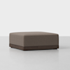A large, square, brown ottoman with a cushioned top and a wooden base, placed on a light, smooth floor against a plain white background.
