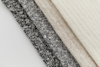 Four fabric samples in various neutral shades, including black, gray, beige, and off-white, are laid out side by side on a white background, showcasing different knit textures and patterns.