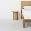 Minimalist bedroom scene featuring a wooden bed frame with a white pillow and white bedding, next to a matching light wood nightstand, set against a plain white wall and light-colored floor.