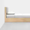 A minimalist wooden bed with a tall headboard featuring a padded gray section, dressed in white bedding and pillow, positioned against a plain white wall on a light gray floor.