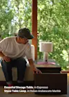 A man wearing a cap and glasses opens a drawer of a dark wood storage table next to a lamp on a side table. Sunlight streams through large windows showing lush green trees outside. Text on image describes the furniture.