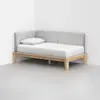 A minimalist daybed with a light wood frame, gray upholstered backrest, and white mattress and pillow, positioned against a white wall in a corner of a room with a gray floor.