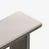 A close-up view of a minimalist bench with a light beige upholstered cushion and a wooden base, set against a plain white background.