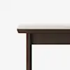 A close-up of a dark wooden bench with a light-colored cushioned seat, shown against a plain white background. Only part of the bench is visible, focusing on one corner and its straight legs.