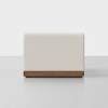 A modern, rectangular ottoman with a light beige upholstered cushion and a wooden base, set against a plain light gray background.