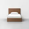 A wooden bed with a rectangular headboard and footboard, neatly made with white bedding and a single white pillow, stands on a light gray floor against a plain white wall.