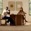 A man and woman sit at a wooden table with cups and plates, facing the camera. Behind them are two abstract art pieces and large windows. The scene is calm and minimalist.