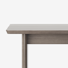 A close-up view of the corner of a wooden table with a light brown finish, showing one leg and part of the tabletop against a plain white background.