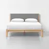 A modern wooden bed frame with a light wood finish, gray upholstered headboard, white bedding, and two white pillows, set in a minimalistic room with a plain white wall and light gray floor.