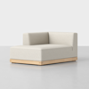 A modern, minimalist corner sofa chair with light beige upholstery and a low, light wood base, set on a smooth, light gray floor against a plain white background.
