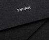 A close-up of black textured material with the word THUMA printed in white, with overlapping layers creating diagonal lines in the background.