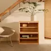 Minimalist bedroom with light wood furniture, open shelves displaying pottery, neutral-toned decor, a beige bed, a textured wall art piece, and exposed wooden ceiling beams above a light rug and smooth floor.