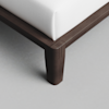 A close-up view of the corner of a dark wooden bed frame with a white mattress on top, highlighting the smooth finish and clean lines of the furniture against a light-colored floor.