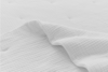 Close-up of a white textured blanket or quilt on a bed, with a gentle wave or fold running through the fabric, creating soft shadows and highlighting the grid-like pattern.