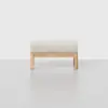 A minimalist wooden stool with a light beige cushioned seat, set against a plain white background.