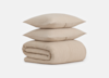 Two beige pillows are stacked on top of a neatly folded beige blanket, all made of a textured fabric, set against a plain white background.