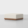 A modern square ottoman with a cream-colored upholstered cushion and a wooden base, set on a smooth, light gray floor against a plain white background.