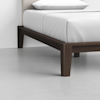 Close-up of the lower corner of a wooden bed frame with dark brown legs and white bedding, set on a light gray floor against a white wall.
