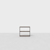 A small, dark wooden shelf with two open shelves stands on a light gray floor against a plain white background.