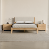 Minimalist wooden bed with light gray upholstered headboard, flanked by matching nightstands. A textured rug covers the concrete floor.