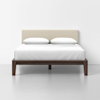 A modern bed with a beige upholstered headboard, white bedding, and 2 white pillows. The bed frame is dark wood, set against a plain white wall and gray floor.