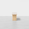 A minimalist wooden bench with a light-colored cushioned seat is centered in a brightly lit, empty space with a smooth, light gray floor and a plain white background.