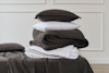 A neatly folded stack of dark gray and white bedding, including pillows, sheets, and blankets, sits on a bed against a white textured wall with decorative molding.