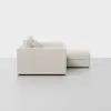Side view of a modern, light beige sectional sofa with three seat cushions and an attached chaise lounge, set against a plain white background.