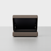 A brown upholstered storage ottoman with its lid open, revealing a spacious black interior compartment. The ottoman sits on a light gray floor against a plain white background.
