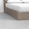Close-up view of the corner of a wooden platform bed with white bedding and pillows, set on a smooth, light gray floor against a plain white wall.