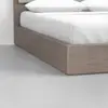 Close-up view of the corner of a wooden platform bed with white bedding and pillows, set on a smooth, light gray floor against a plain white wall.