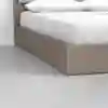 Close-up view of the corner of a wooden platform bed with white bedding and pillows, set on a smooth, light gray floor against a plain white wall.