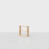 A small, light wood, three-tier shelf stands alone on a gray, textured floor against a plain white background. The design is minimal and modern, with open sides and clean lines.