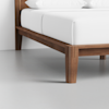 Close-up of a wooden bed frame with tapered legs on a light gray floor, showing white bedding and pillows. The image highlights the corner and construction details of the bed.