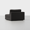 A modern, dark gray armless sectional sofa chair is shown at an angle on a light gray floor with a plain, light background. The upholstery is clean and minimalistic, featuring straight lines and a boxy design.