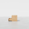 A light wood two-drawer cabinet stands on a smooth gray floor against a plain white background, with the bottom drawer partially open.