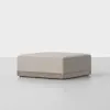 A square, beige upholstered ottoman with a wooden base sits on a smooth, light gray floor against a plain white background.