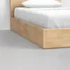 A close-up of a wooden bed frame with white bedding and a pillow, set on a smooth, light gray floor against a plain white wall.