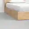 A close-up of a wooden bed frame with white bedding and a pillow, set on a smooth, light gray floor against a plain white wall.