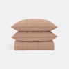 Two light brown pillows are stacked on top of a matching folded quilt, all featuring a subtle grid stitching pattern, against a plain white background.