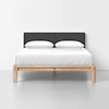 A modern bed with a light wood frame, a dark gray upholstered headboard, white bedding, and two white pillows, set against a plain white wall on a smooth, light gray floor.