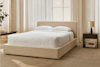 A neatly made bed with white linens and two pillows sits in a bright, minimalist bedroom with beige carpet, a nightstand, a lamp, and large windows with sheer curtains letting in natural light.