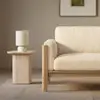 A beige sofa with wooden frame sits next to a small stone side table with a cream-colored lamp, set against a neutral wall with framed art. The decor is minimal and modern.