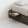 A minimalist wooden bedside tray holds a black coffee cup, saucer, and a closed notebook with a pen, attached to a dark wood bed frame with beige bedding.