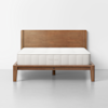 A minimalist wooden bed frame with a rectangular headboard and a white mattress sits against a plain white wall on a light gray floor. The bed frame has clean lines and the word “THUMA” is stitched on the mattress.