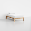 A minimalist single bed with a light wooden frame, white mattress, and a white pillow, set against a plain white wall and floor in a simple, modern room.