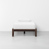 A single bed with a dark wooden frame, neatly made with white bedding and a white pillow, positioned against a plain white wall in a minimalistic room with a light gray floor.