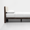 A minimalist wooden bed frame with a beige upholstered headboard, white sheets, blanket, and pillow, positioned against a plain white wall on a light-colored floor.