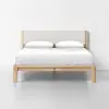 A modern wooden bed frame with a light beige upholstered headboard, neatly made with white bedding and two pillows, set against a plain white wall and floor.