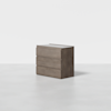 A modern, three-drawer wooden dresser with a light brown finish, placed on a smooth, light gray floor against a plain white background.